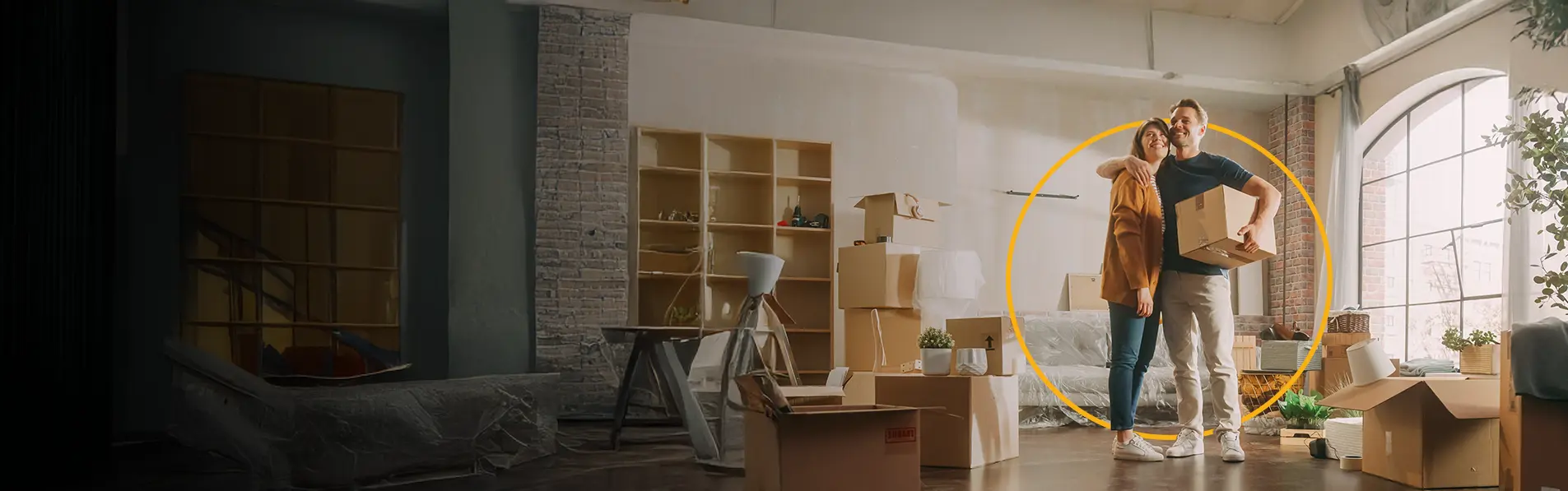 A couple hugging eachother with boxes and packed furniture surrounding their home.