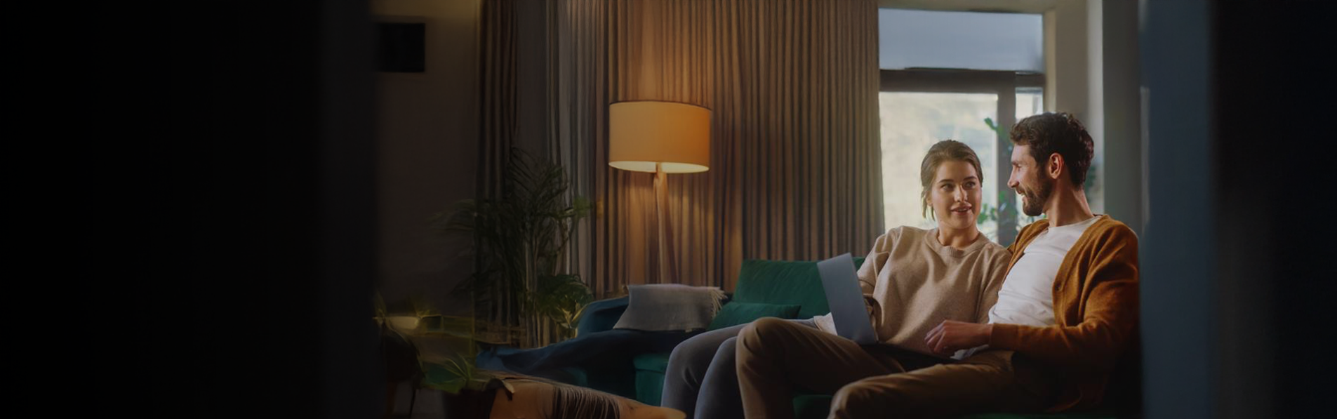 A couple sat on a couch with a laptop on their knees talking.