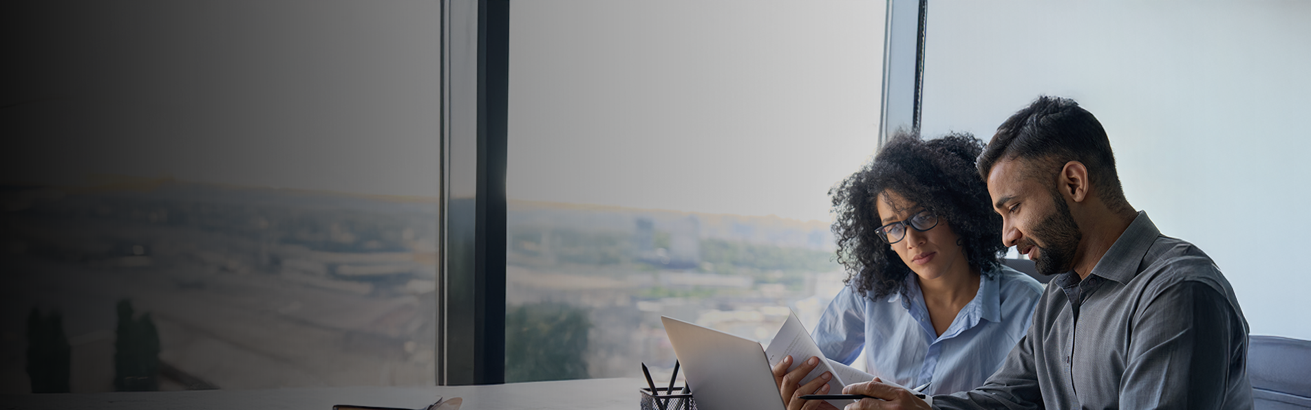 A man and a woman sat down looking at a laptop together in a high rise office.