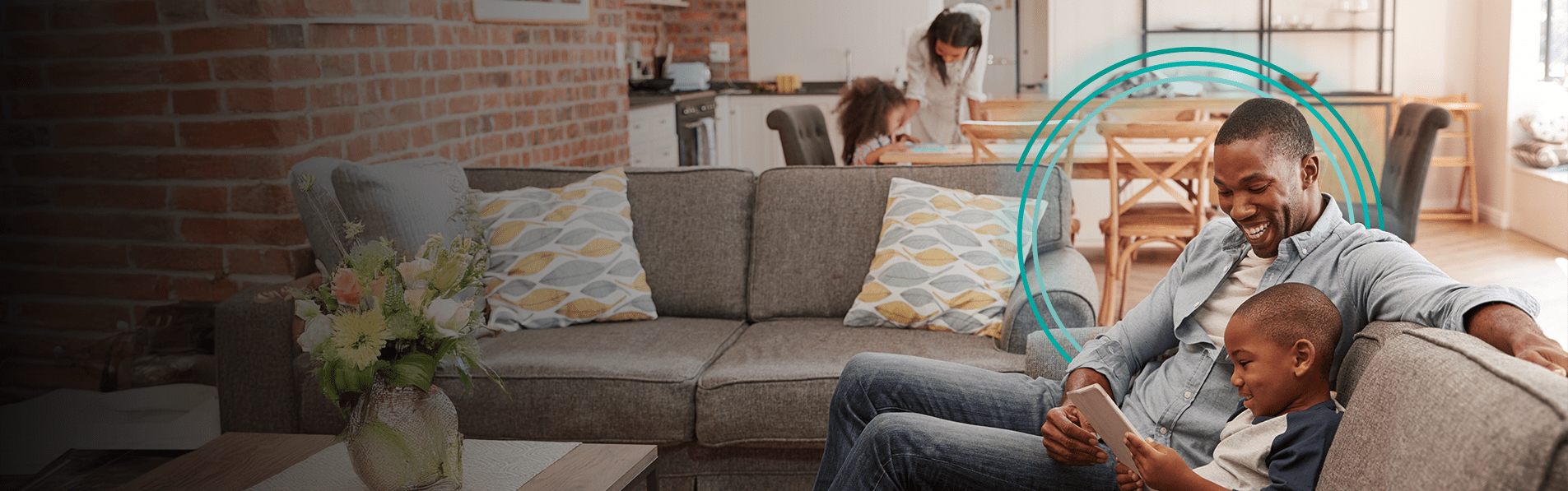 A man and his son sat on the couch with an iPad whilst his wife and daughter play in the background.