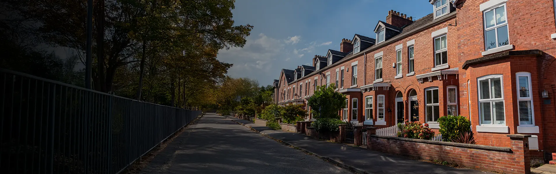 A row of red brick terraced houses on a street opposite a fenced off park.