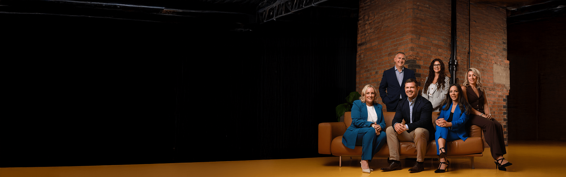 A wide shot of the senior leadership team sitting on a leather couch in front of a red brick wall.