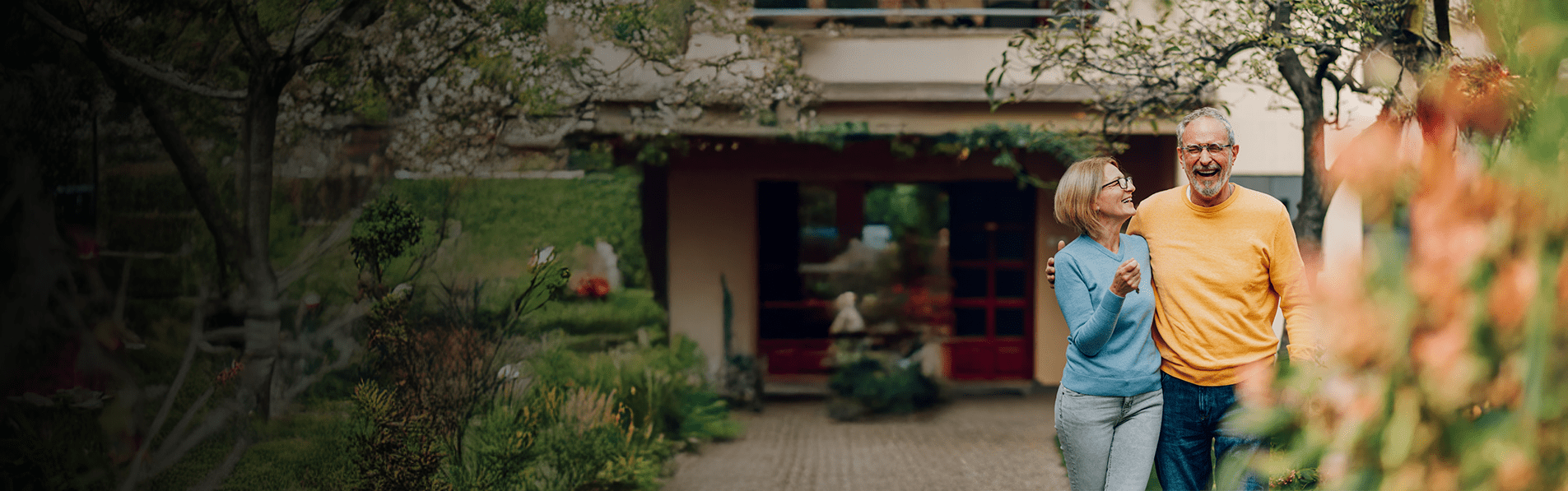 An elderly couple walking out of their house laughing with their arms around each other.