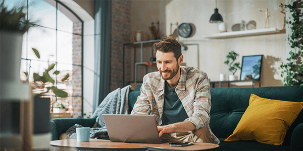 A man sat on a couch smiling whilst on his laptop in a modern styled house.