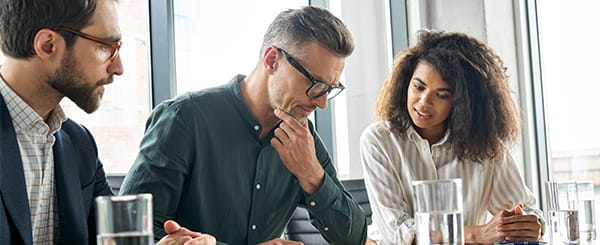 Two men in suits and glasses sat with a woman looking at documents