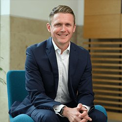 Andrew Hankin with short blonde hair smiling sat on a chair.