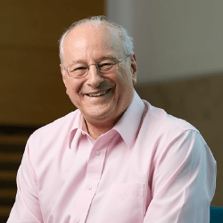 An older professional male with short grey hair and glasses smiling wearing a pink shirt.