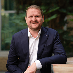 Ryan Etchells wearing a blue navy suit smiling sat on a chair.