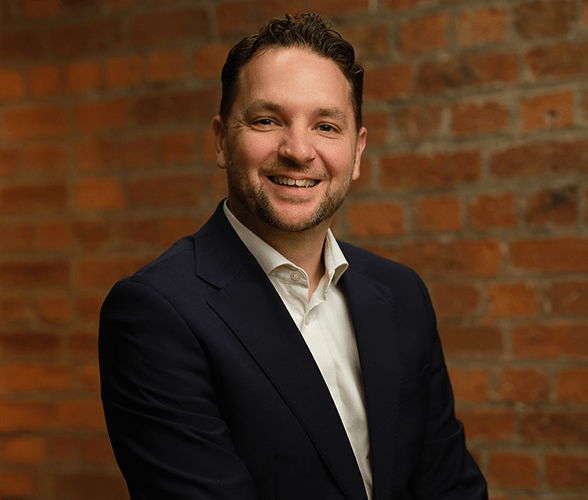 Andrew Douglas smiling with short brown hair in front of a red brick wall.
