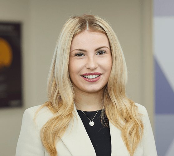 A woman with long blonde hair smiling wearing a beige blazer and a black top.