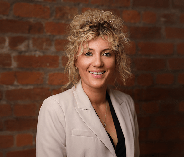 Kara Williams with long curly blonde hair smiling in front of a red brick wall.