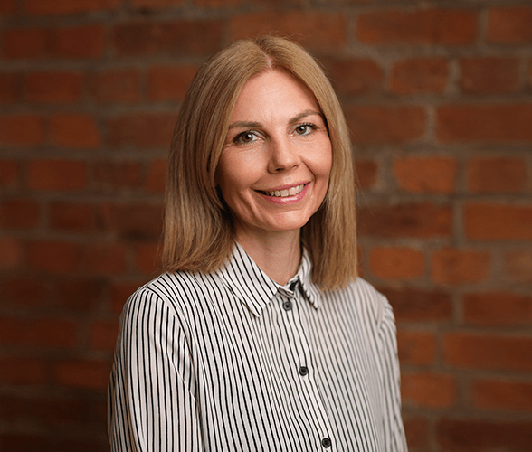 Katie Wilsher-Garratt with long blonde hair smiling in front of a red brick wall.