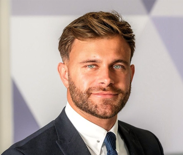 A man with short light brown hair smiling wearing a suit.