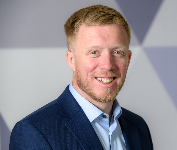 A man with short ginger hair and a beard smiling wearing a navy blue blazer and light blue shirt.