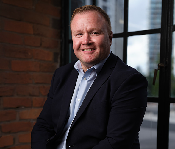 A man with short ginger hair smiling wearing a black blazer and blue shirt.