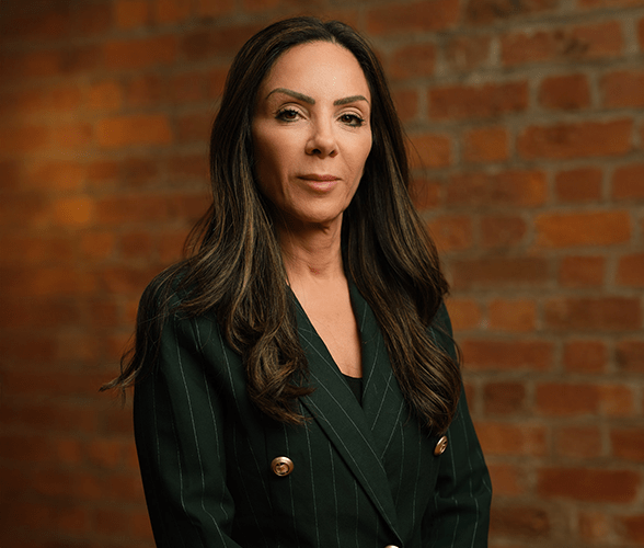Tanya Elmaz with long brown hair smiling in front of a red brick wall.