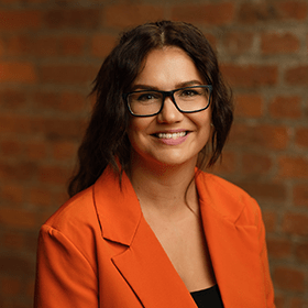 A woman with long brown hair tied up smiling wearing an orange blazer and black top.