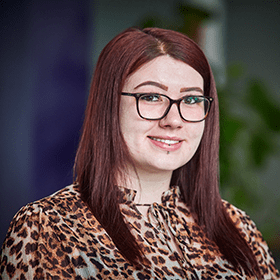 A woman with medium length red hair smiling wearing glasses and a cheetah print blouse.