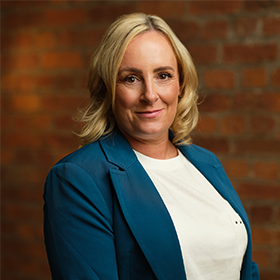 A woman with short blonde hair smiling wearing a teal blazer and a white shirt.