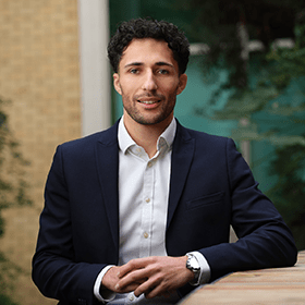 A man with short curly black hair smiling wearing a suit.