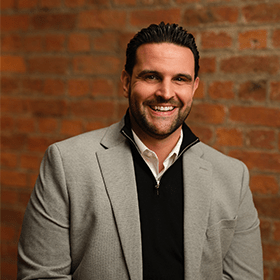 Dan Narwal with short black hair smiling in front of a red brick wall.