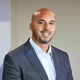 A bald man with a black beard smiling wearing a grey suit and white shirt.