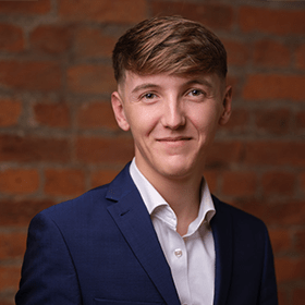 Freddie Atkinson with short light brown hair smiling in front of a red brick wall.