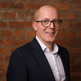 Gareth Wilde smiling in front of a red brick wall.