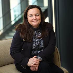 A mid-shot of Helen Calderley with long brown hair smiling wearing a black jacket with a blouse underneath.