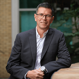 A man with short grey hair smiling with glasses on wearing a suit.