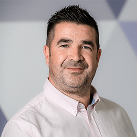 A man with short black hair and a stubble smiling, wearing a light pink shirt.