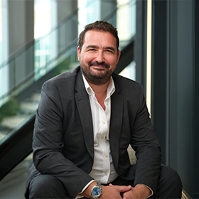 A mid-shot of James Steadman with short brown hair and a beard smiling wearing a smart suit.