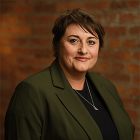 Jo Elton with short brown hair smiling in front of a red brick wall.