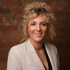 Kara Williams with long curly blonde hair smiling in front of a red brick wall.