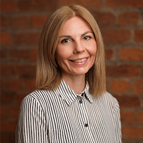 Katie Wilsher-Garratt with long blonde hair smiling in front of a red brick wall.