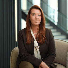 A mid-shot of Kristi Capaldi with long ginger hair smiling wearing a brown jacket and a blouse underneath.
