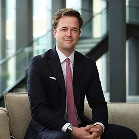 A mid-shot of Lewis Hepworth with short brown hair smiling wearing a smart suit.