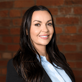 Lois Ashcroft with long brown hair smiling in front of a red brick wall.