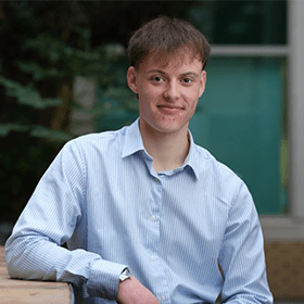 A young man with short brown hair smiling wearing a light blue shirt.