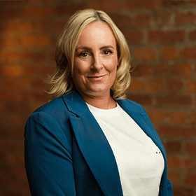 Maeve Ward with short blonde hair smiling in front of a red brick wall.