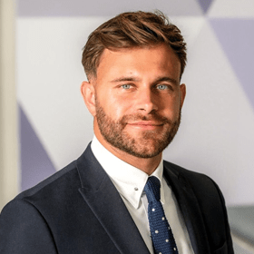 A man with short light brown hair smiling wearing a suit.