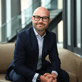 A mid-shot of Phil Ryan wearing glasses and smiling with a smart suit on.