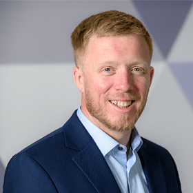 A man with short ginger hair and a beard smiling wearing a navy blue blazer and light blue shirt.