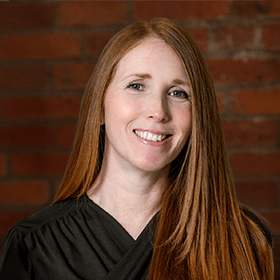 Sadie Harrison with long ginger hair smiling in front of a red brick wall.