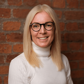 Sally Precious Ward with medium length blonde hair smiling in front of a red brick wall.