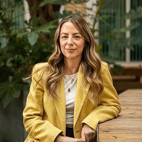 A mid-shot of Stacey Doyle with long brown hair, a white top and a yellow blazer smiling.