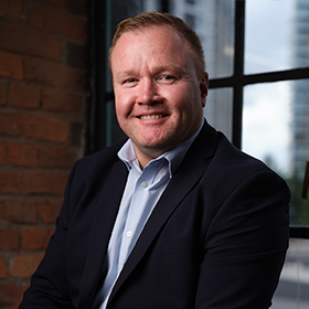 A man with short ginger hair smiling wearing a black blazer and blue shirt.