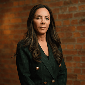 Tanya Elmaz with long brown hair smiling in front of a red brick wall.