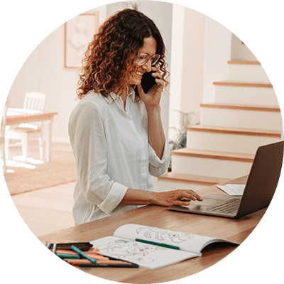 A close up of a woman sat at a desk on the phone whilst typing on a laptop.