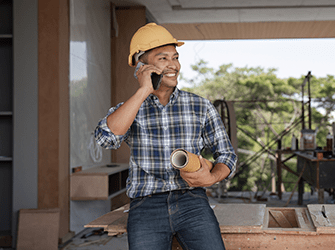 A man wearing a hardhat talking on the phone with blueprints in his hand.